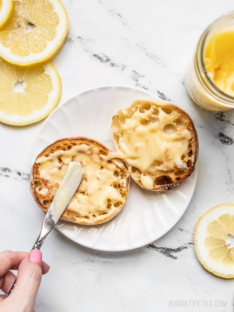 Lemon curd being spread onto a toasted english muffin, with the jar of curd and lemon slices on the sides.