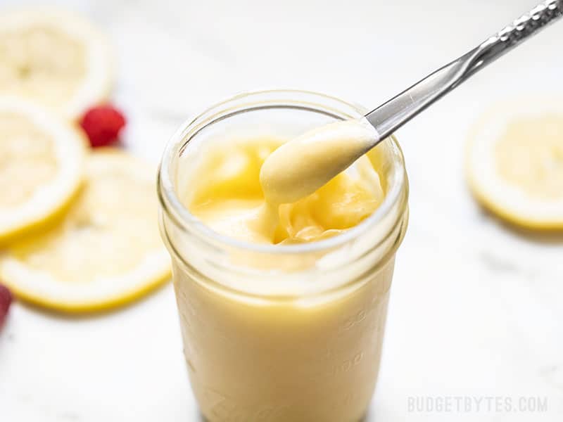 This luscious homemade lemon curd only takes four simple ingredients, about 10 minutes, and tastes like velvet sunshine in a jar. Budgetbytes.com Homemade lemon curd being lifted out of a jar with a small silver butter knife, lemon slices in the background.