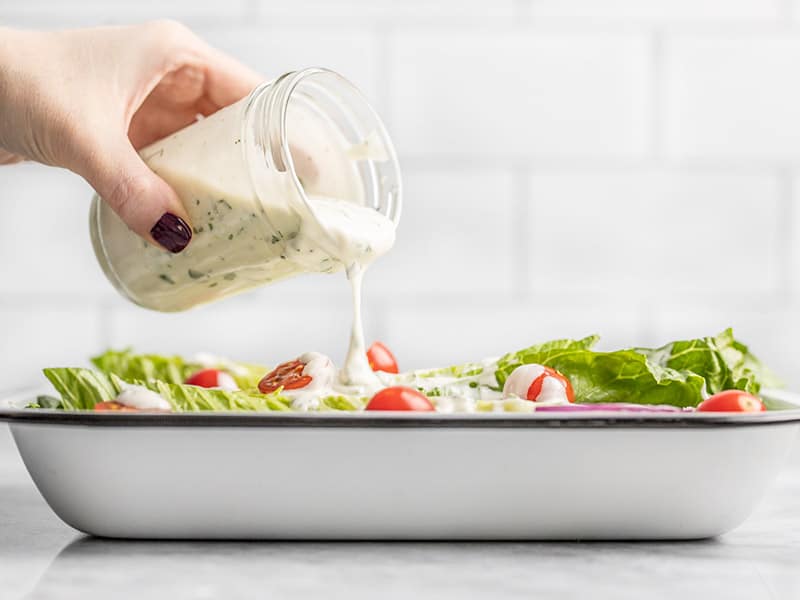 Homemade ranch dressing is creamy, tangy, and fully of savory herbs and spices. Drizzle on your favorite salad, or use as a dip for vegetables and chips! Budgetbytes.com Homemade Ranch Dressing Being Poured onto a salad from a jar, viewed from the side.