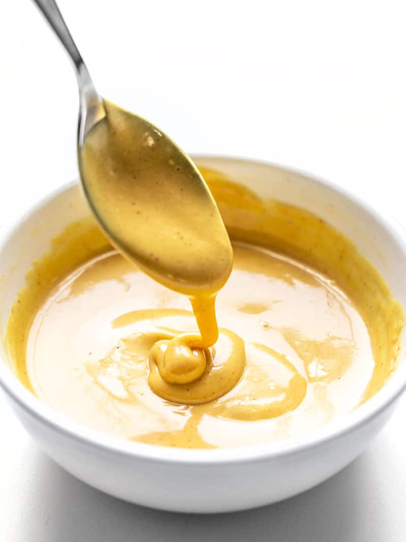This sweet, tangy, and creamy homemade honey mustard sauce only takes a few minutes to make and is great for sandwiches, salads, dipping, and more! Budgetbytes.com Honey Mustard Sauce dripping off a spoon into a white bowl.