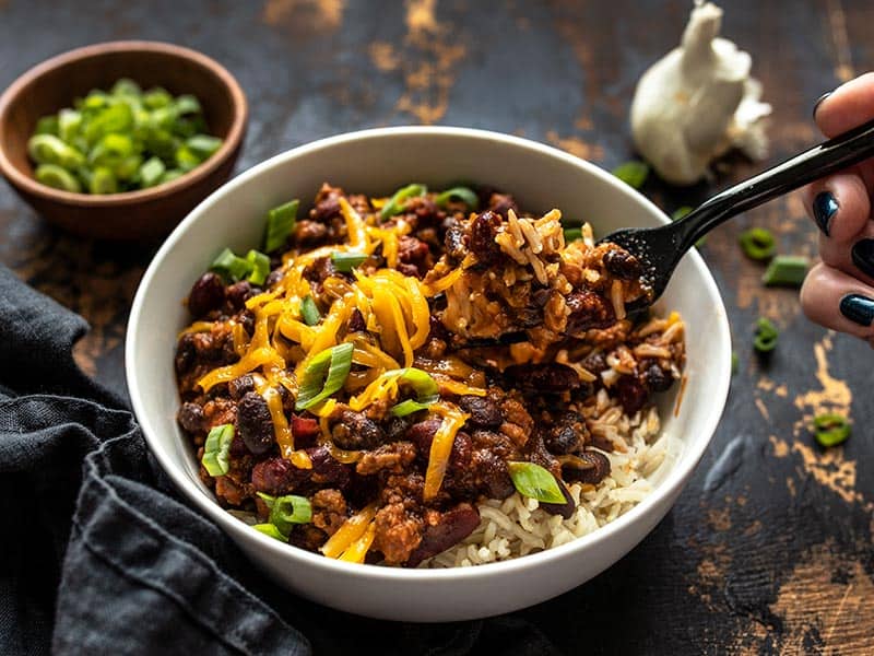 Serve these sweet, tangy, and savory BBQ Beef and Beans with rice and cheese for a simple and comforting weeknight dinner the whole family will love. Budgetbytes.com Front view of a bowl of BBQ Beef and Beans topped with cheddar and green onion, and a fork lifting a bite.