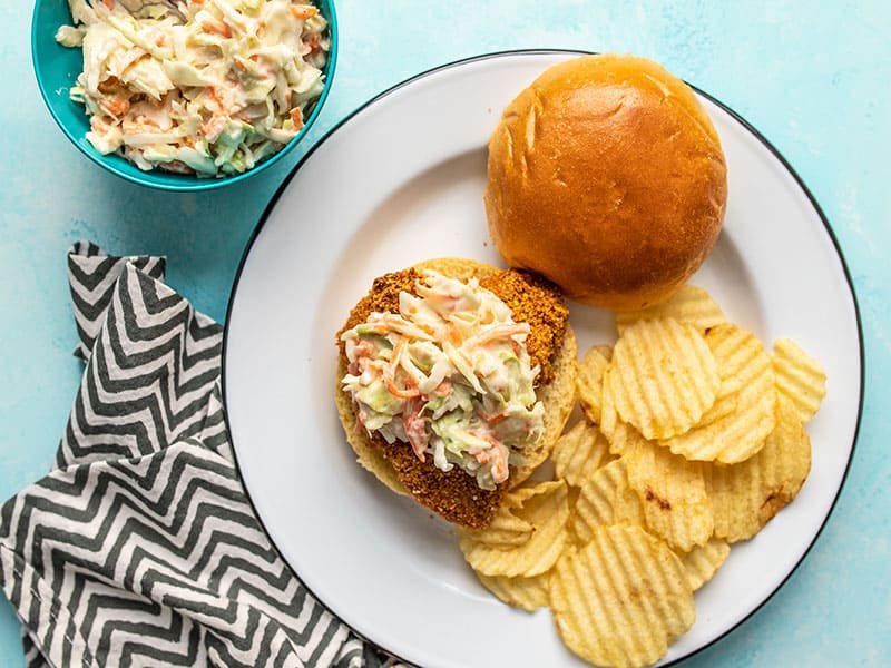 These baked spicy chicken sandwiches have three layers of heat and are topped with a creamy-sweet quick homemade coleslaw. Budgetbytes.com One Baked Spicy Chicken Sandwich on a plate with wavy potato chips and a bowl of honey yogurt slaw on the side.