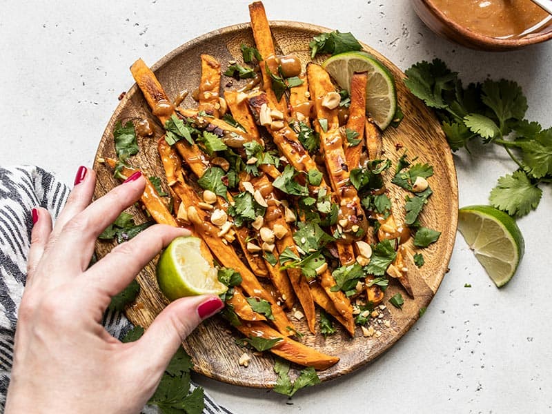 Oven baked sweet potato fries drizzled with homemade peanut lime dressing, piled high with fresh cilantro, chopped peanuts, and a pinch of spicy red pepper. Budgetbytes.com A wedge of lime being squeezed over a plate of baked sweet potato fries with peanut lime dressing.