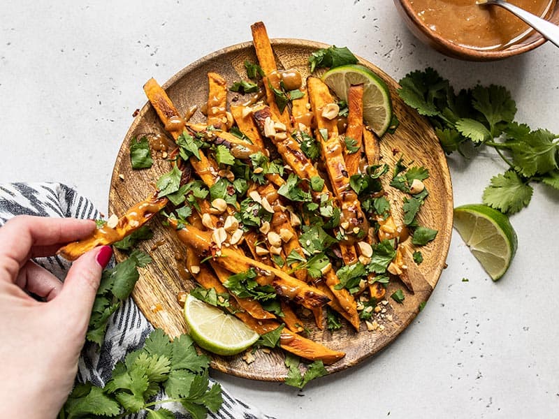 Oven baked sweet potato fries drizzled with homemade peanut lime dressing, piled high with fresh cilantro, chopped peanuts, and a pinch of spicy red pepper. Budgetbytes.com A plate full of baked sweet potato fries and peanut lime dressing, a hand sneaking one fry off the side.