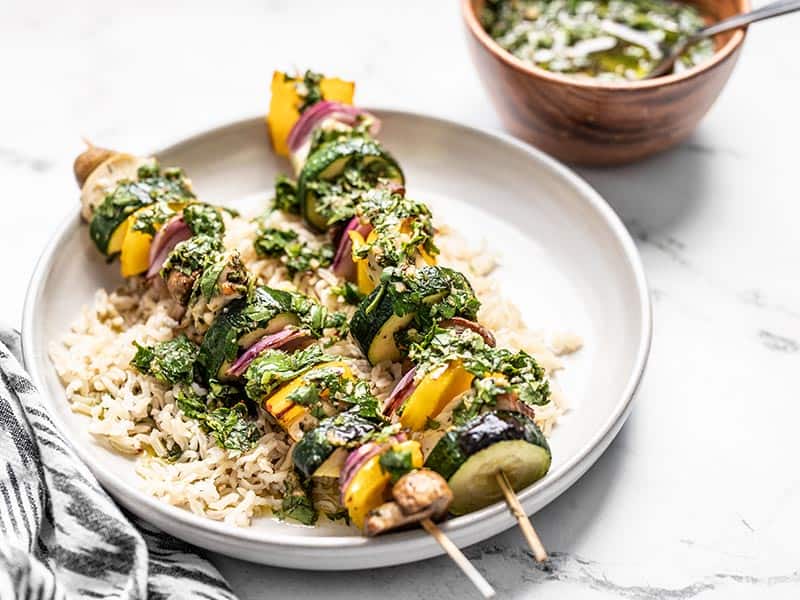 These Chimichurri Chicken Kebabs feature fresh vegetables and marinated chicken cooked over an open flame, then drizzled with a vibrant chimichurri sauce. Budgetbytes.com Front view of a plate with rice and two chimichurri chicken kebabs, with a wooden bowl of chimichurri in the back.