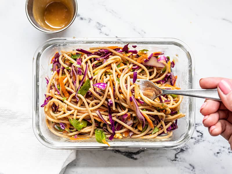 This Cold Peanut Noodle Salad with homemade peanut lime dressing is the perfect meal prep for summer! No reheating necessary. Budgetbytes.com A meal prep container of Cold Peanut Noodle Salad being eaten with a fork, the empty dressing container on the side.