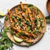 A wooden plate full of baked sweet potato fries drizzled with peanut lime dressing and topped with cilantro, peanuts, and red pepper.