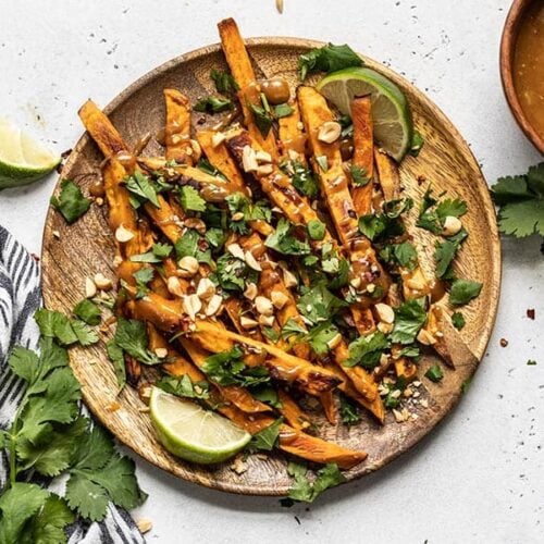 A wooden plate full of baked sweet potato fries drizzled with peanut lime dressing and topped with cilantro, peanuts, and red pepper.
