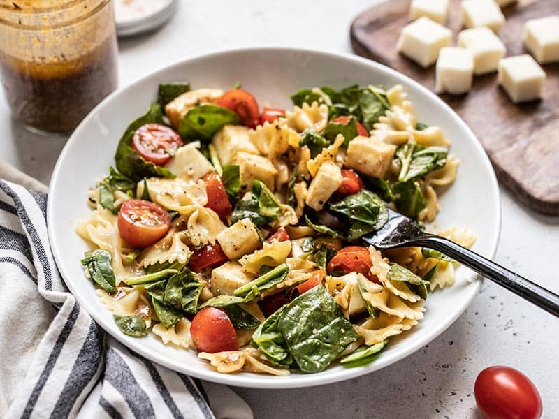 This Tomato Mozzarella Pasta Salad with homemade Parmesan Balsamic Vinaigrette is the perfect light and fresh summer meal. Perfect for potlucks! Budgetbytes.com Front view of a bowl of Tomato Mozzarella Salad with a jar of dressing in the back and cubed mozzarella on the side.