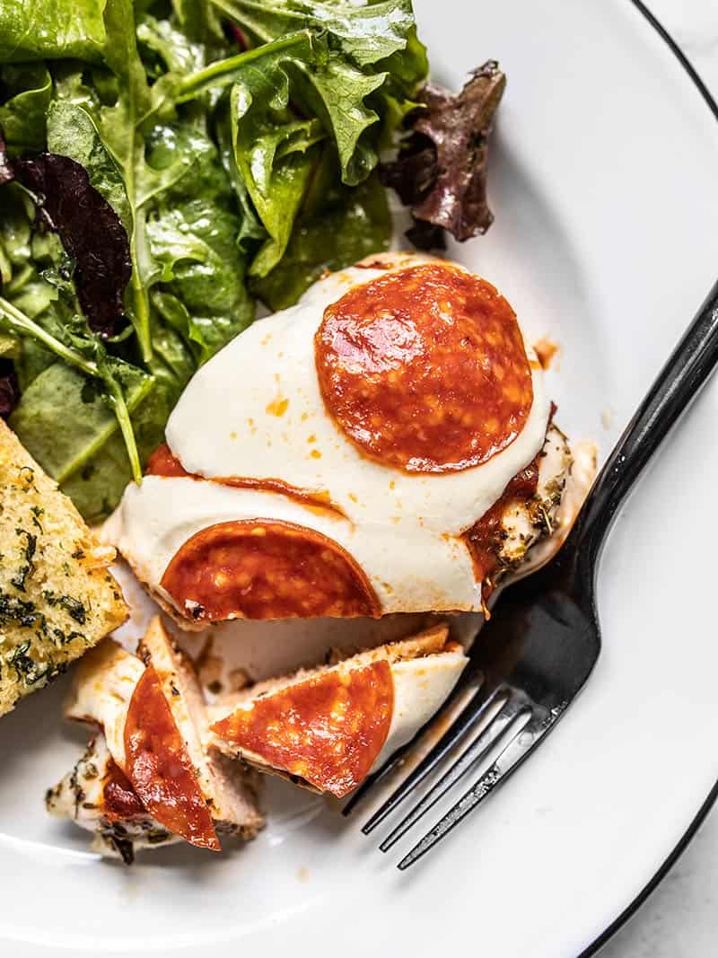 This easy Baked Pizza Chicken is the perfect fast and easy weeknight dinner. The chicken cooks quickly and always stays tender and juicy! BudgetBytes.com Close up of Baked Pizza Chicken on a plate with salad and garlic bread, the chicken cut into pieces.