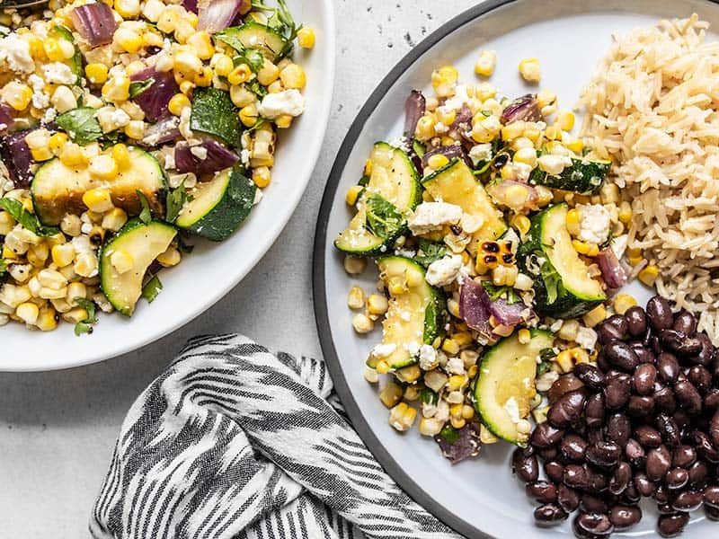 This Charred Corn and Zucchini Salad perfectly captures the flavor of summer with fresh sweet corn, salty feta, and a smoky grill-like flavor. BudgetBytes.com A plate filled with Charred Corn and Zucchini Salad, black beans, and rice, next to a bowl of the corn and zucchini salad.