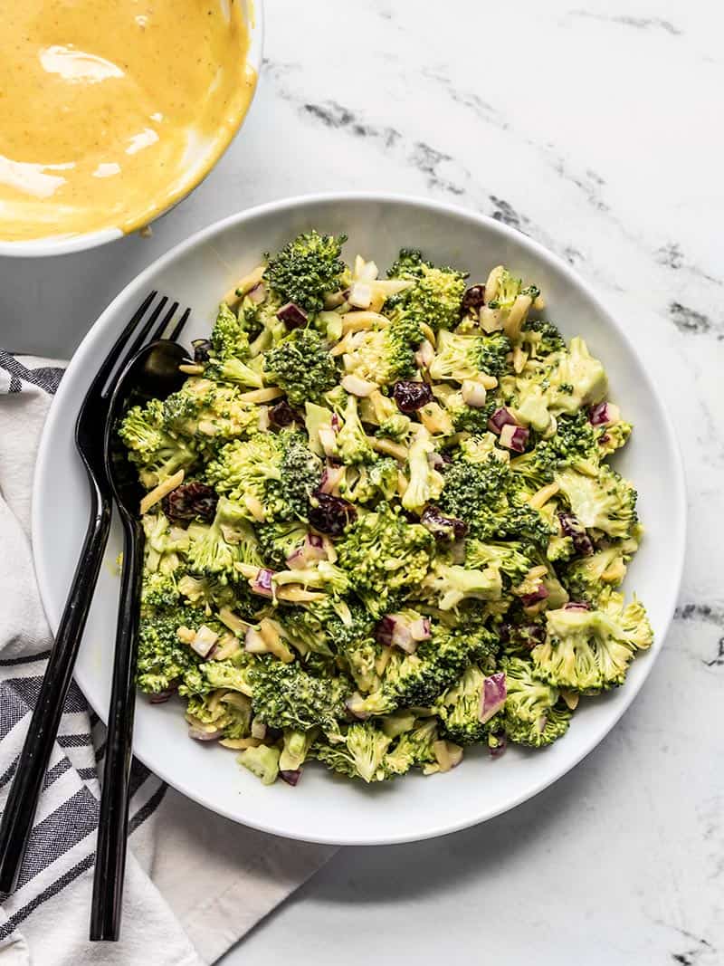 A bowl of honey mustard broccoli salad with black utensils on the side of the bowl and a bowl of honey mustard sauce near by.