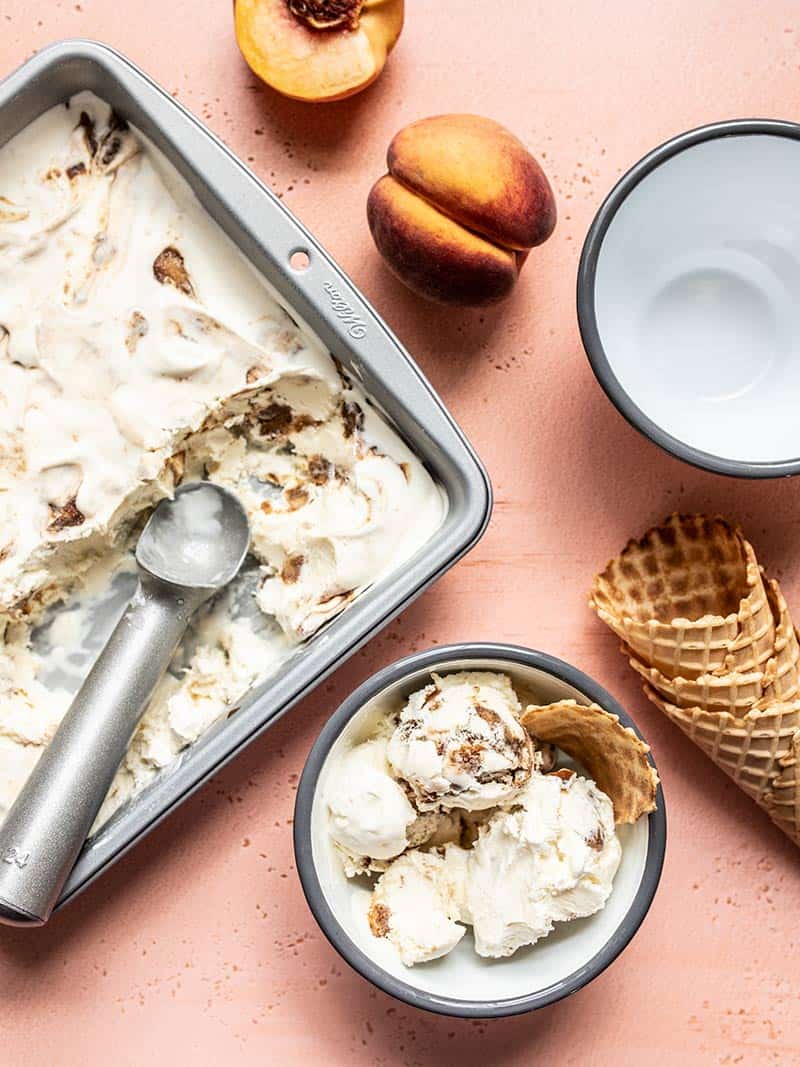 This No Churn Balsamic Peach Ice Cream is incredibly easy and pairs the flavor of fresh sweet summer peaches with a tangy and buttery balsamic glaze. BudgetBytes.com No Churn Balsamic Peach Ice Cream scooped out of the freezer dish into one out of two bowls, waffle cones on the side.