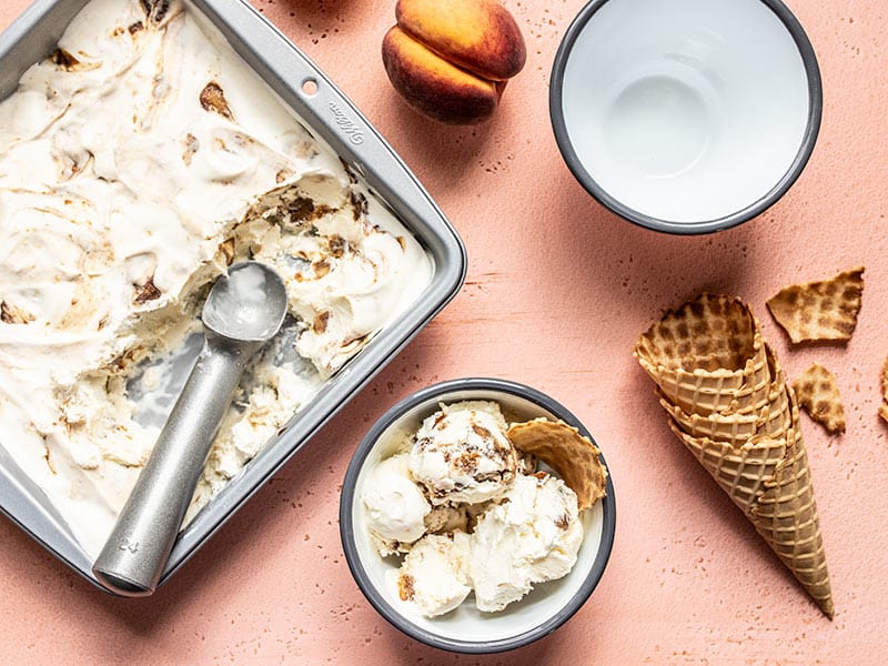 This No Churn Balsamic Peach Ice Cream is incredibly easy and pairs the flavor of fresh sweet summer peaches with a tangy and buttery balsamic glaze. BudgetBytes.com Baking pan full of No Churn Balsamic Peach Ice Cream scooped out and served in one bowl, with one empty bowl, and some waffle cones.