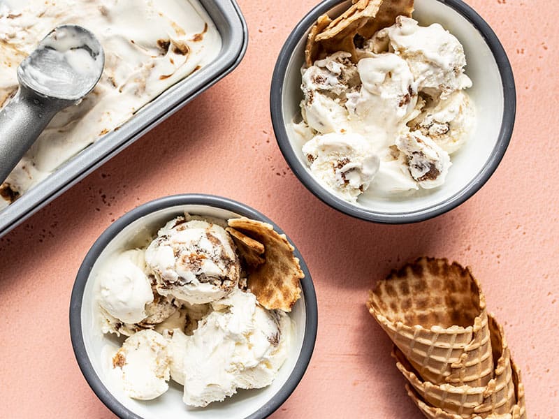 This No Churn Balsamic Peach Ice Cream is incredibly easy and pairs the flavor of fresh sweet summer peaches with a tangy and buttery balsamic glaze. BudgetBytes.com Two bowls of No Churn Balsamic Peach Ice Cream with the pan of ice cream and a stack of waffle cones on the side.
