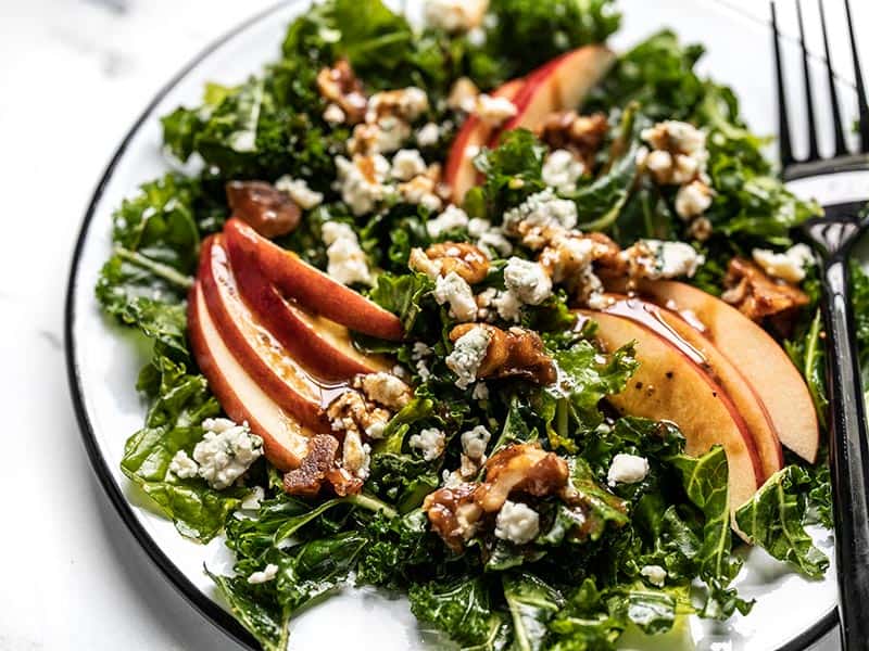 All the flavors of fall come together in this Autumn Kale and Apple Salad, with candied walnuts and a homemade balsamic vinaigrette. BudgetBytes.com Close up side view of Autumn Kale and Apple Salad on the plate with dressing.