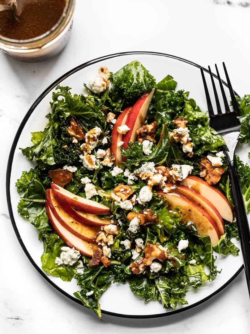 All the flavors of fall come together in this Autumn Kale and Apple Salad, with candied walnuts and a homemade balsamic vinaigrette. Overhead view of a plate full of Autumn Kale and Apple Salad, drizzled with balsamic vinaigrette and a black fork on the side of the plate.