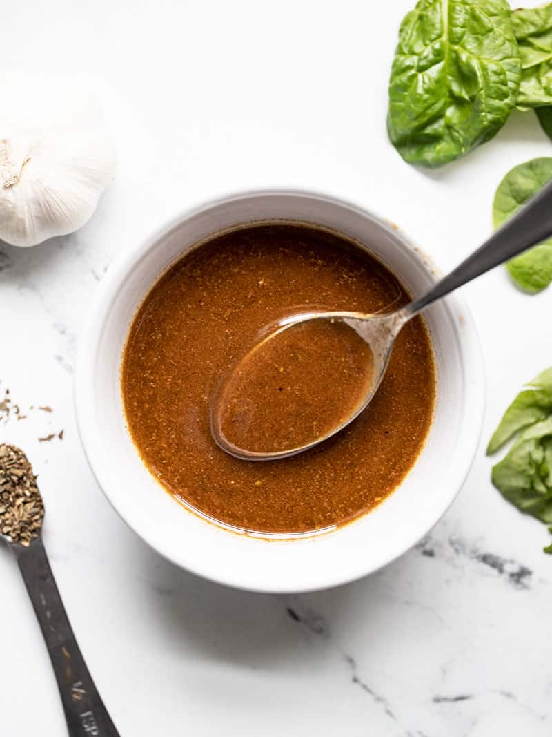 This super simple homemade balsamic vinaigrette is perfect for all your green salads and pasta salads and has several options for customizing! BudgetBytes.com A small white bowl of homemade balsamic vinaigrette with a spoon in it and spinach, garlic, and dried basil on the side