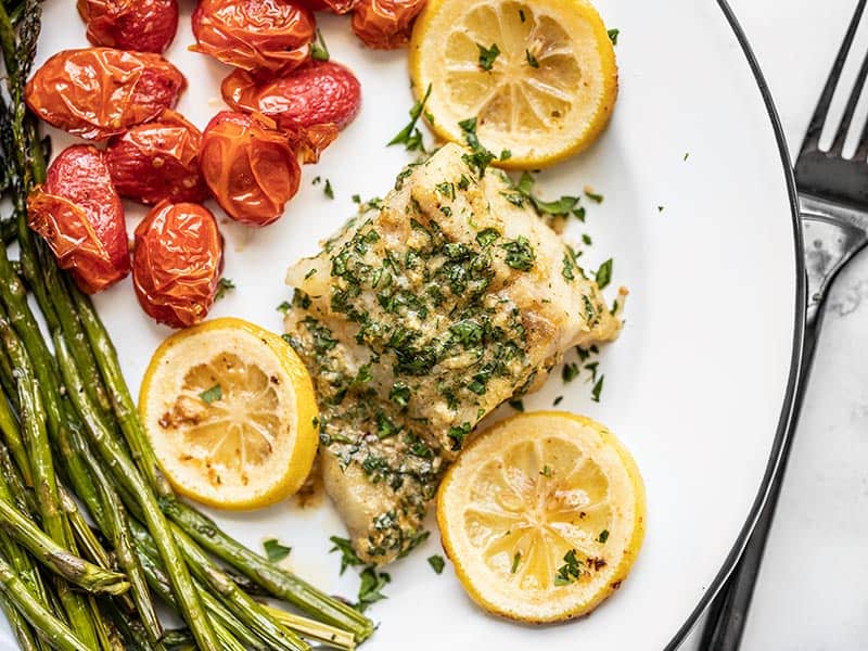 This rich and zesty Garlic Butter Baked Cod is a fast and flavorful weeknight dinner that can be made in under 30 minutes, using basic pantry staples.BudgetBytes.com Overhead view of Garlic Butter Baked Cod on a plate with tomatoes and asparagus, a black fork on the side.