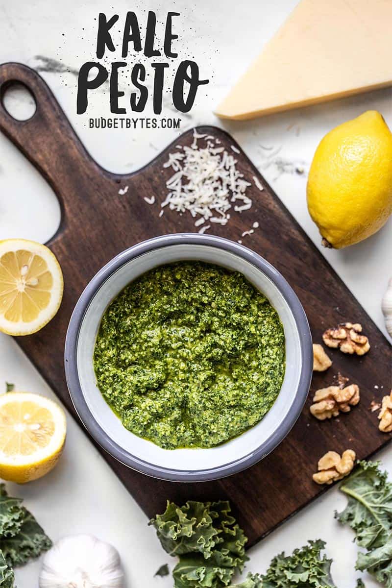 Kale pesto in a bowl on a cutting board with ingredients scattered all around.