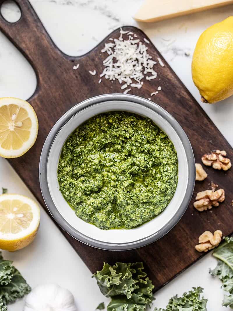 Making a small batch of kale pesto is a great way to use leftover kale. This flavorful pesto is great on sandwiches, in soups, dips, and more! A bowl of Kale Pesto on a wooden cutting board surrounded by lemons, walnuts, kale, and Parmesan