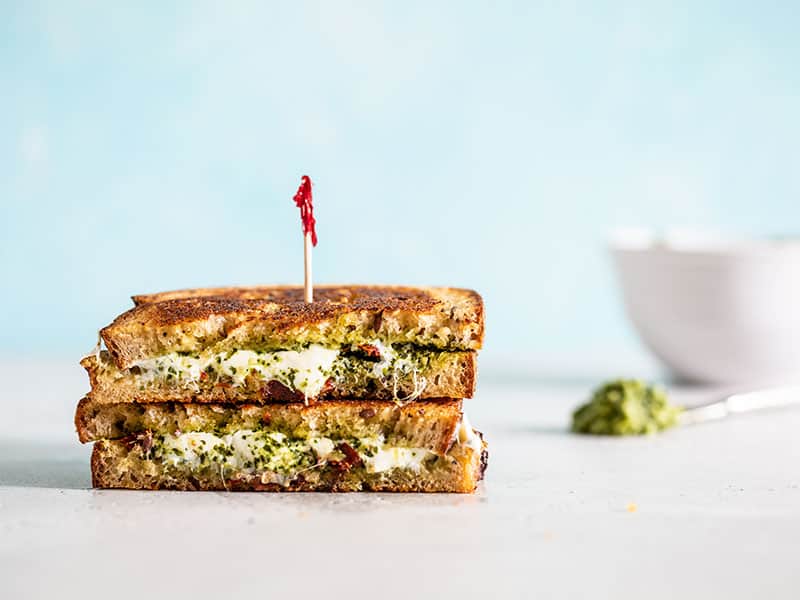 Whether you're using up leftover pesto or just want to treat yourself to something special, this Pesto Grilled Cheese is next level! BudgetBytes.com Two stacked pieces of pesto grilled cheese next to a bowl and small knife with pesto on it.