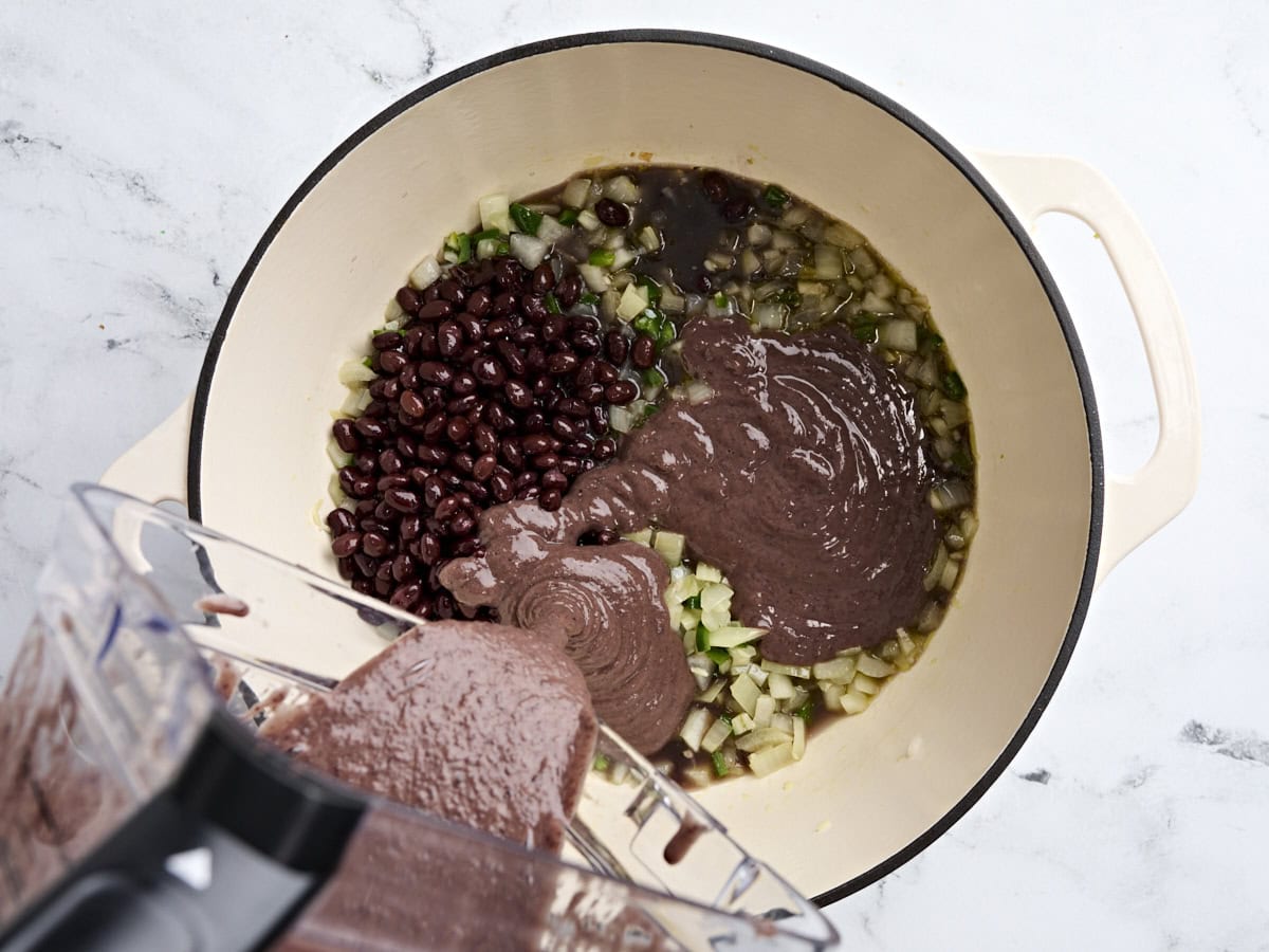 Pureed black beans being poured into a soup pot with diced veggies in.