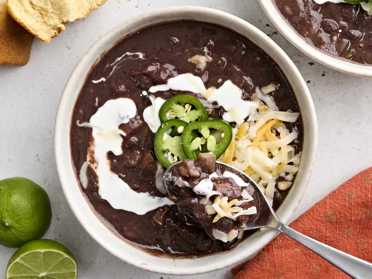 Overhead view of a spoon taking some black bean soup from a bowl, which is topped with jalapeno slices, shredded cheese, and sour cream.