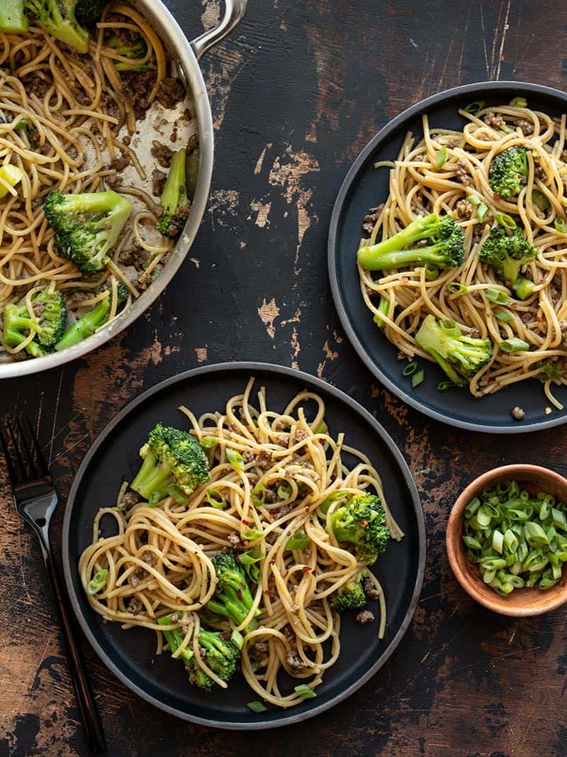 Garlic Noodles with Beef and Broccoli is a quick and easy all-in-one bowl dinner recipe with a rich, buttery, salty, and sweet sauce. BudgetBytes.com Two black plates of Garlic Noodles with Beef and broccoli next to the pan and a bowl of sliced green onions.