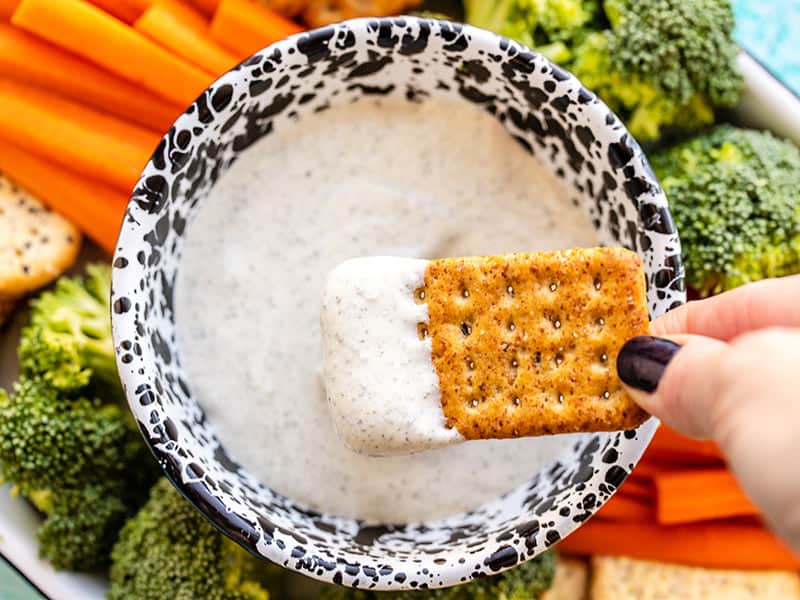 Say goodbye to curds because this Garlic Herb Cottage Cheese Dip is whipped until smooth and creamy for a high protein vegetable and chip dip! BudgetBytes.com A cracker being dipped into the whipped cottage cheese dip, the platter of vegetables and crackers in the background