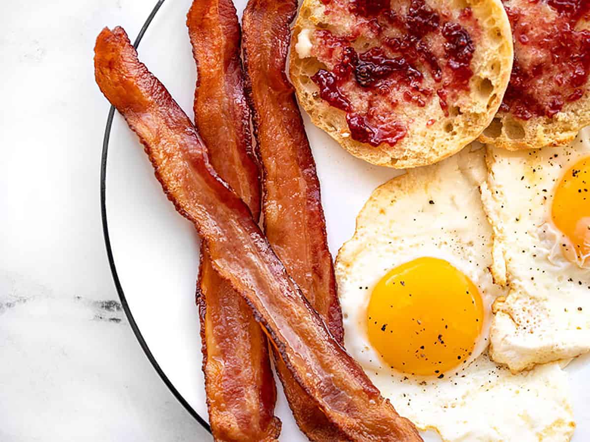 Learn how to cook bacon in the oven with very little effort and no hot splattering bacon grease! Perfect, crispy bacon in the oven every time. BudgetBytes.com Close up of a plate with oven baked bacon, and egg, and English muffin.