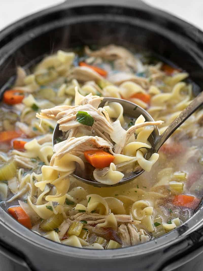 Slow cooker chicken noodle soup is an incredibly easy and soothing winter recipe that will fill you up and warm you from the inside out! BudgetBytes.com Front view of a ladle lifting some chicken noodle soup out of the slow cooker