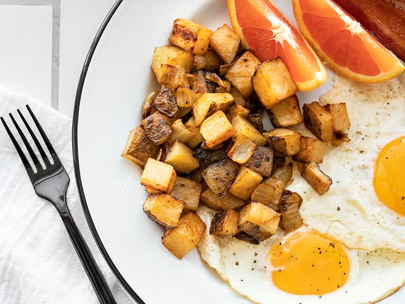 These Smoky Roasted Breakfast Potatoes cook in the oven, leaving your hands and pans free to cook your eggs and enjoy a stress-free breakfast! BudgetBytes.com Close up of roasted breakfast potatoes on a plate with eggs and orange slices