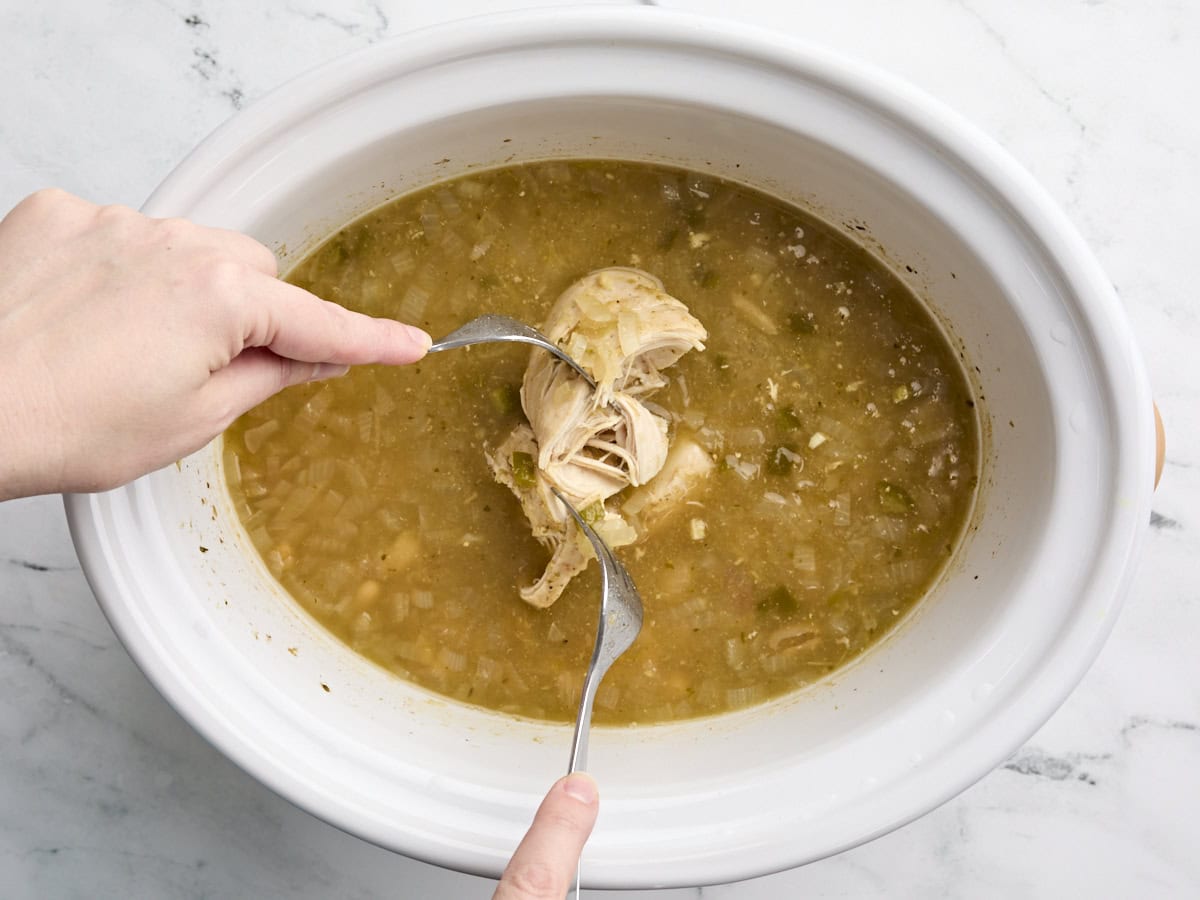Hands using two forks to shred a chicken breast in a crockpot.