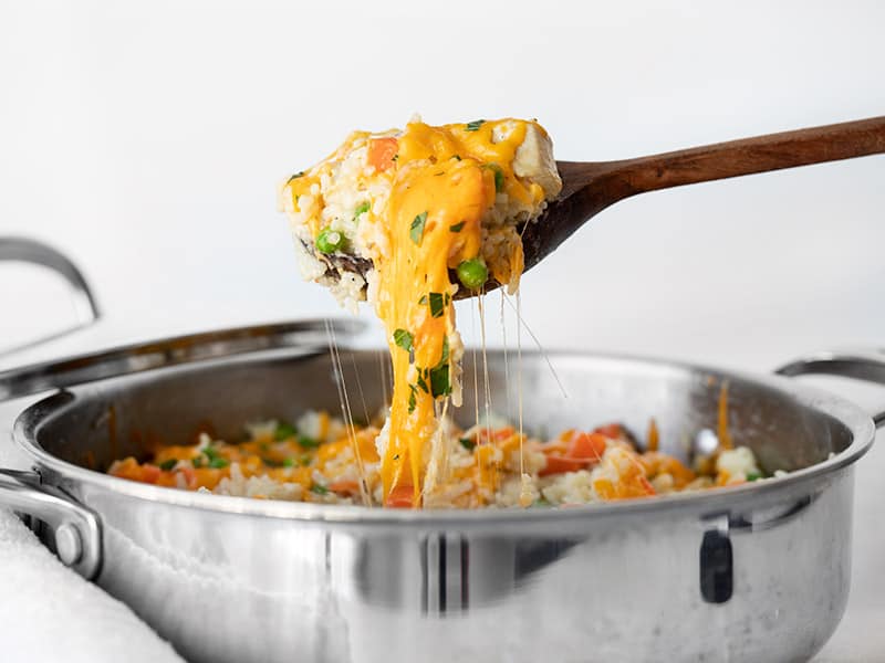 This easy one pot Creamy Chicken and Rice Skillet makes a comforting weeknight dinner during those long, dark winter months! Makes great leftovers, too! BudgetBytes.com A wooden spoon lifting chicken and rice out of the skillet with melted cheese stretching between