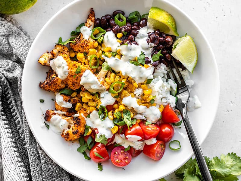 These light and flavorful Roasted Cauliflower Taco Bowls have tons of color, flavor, and texture to keep you coming back for more! Great for meal prep! BudgetBytes.com Close up overhead view of a roasted cauliflower taco bowl topped with cilantro lime ranch and a black fork in the middle