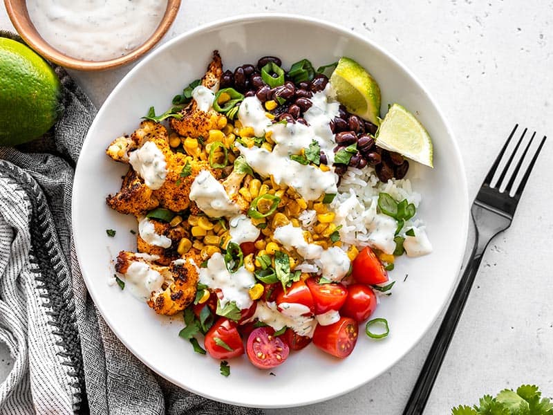 These light and flavorful Roasted Cauliflower Taco Bowls have tons of color, flavor, and texture to keep you coming back for more! Great for meal prep! BudgetBytes.com A finished roasted cauliflower taco bowl with cilantro lime ranch, with a black fork on the side.
