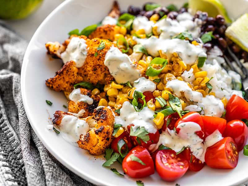 These light and flavorful Roasted Cauliflower Taco Bowls have tons of color, flavor, and texture to keep you coming back for more! Great for meal prep! BudgetBytes.com Close up side view of a roasted cauliflower taco bowl topped with cilantro lime ranch