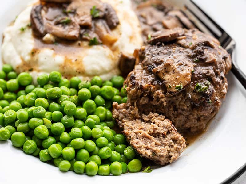 Homemade Salisbury Steak with a rich mushroom gravy is a quick, simple, and hearty weeknight dinner that will fill even the biggest of appetites! BudgetBytes.com Close up side view of a Salisbury Steak on a plate with peas and mashed potatoes, and a piece of the steak cut off the side.