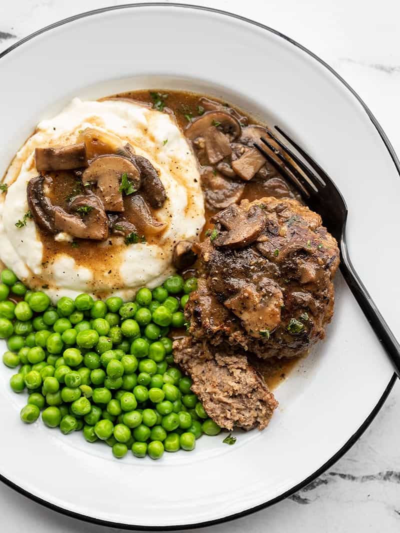 Homemade Salisbury Steak with a rich mushroom gravy is a quick, simple, and hearty weeknight dinner that will fill even the biggest of appetites! BudgetBytes.com Salisbury steak with mushroom gravy on a plate with mashed potatoes and peas, a fork on the side.