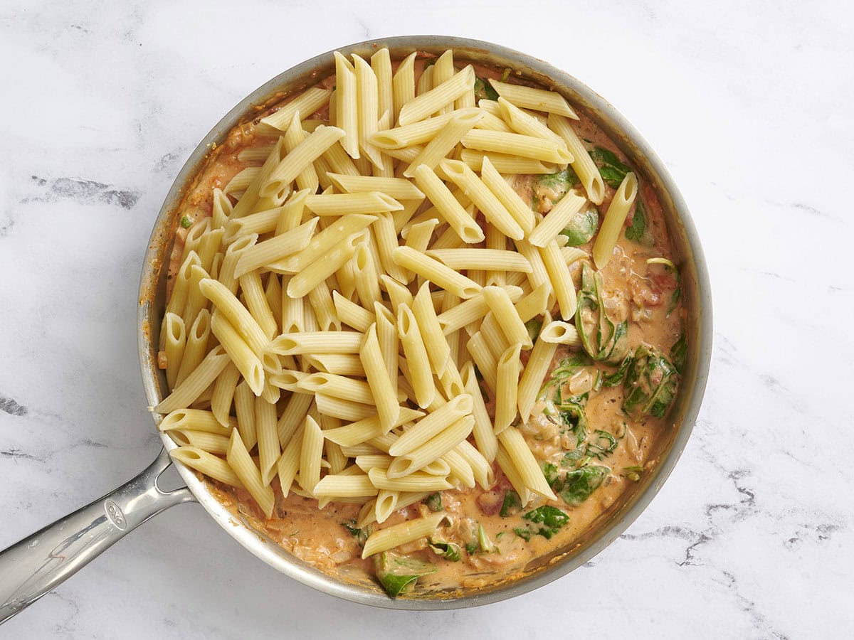 Cooked pasta added to creamy tomato sauce with spinach in a skillet.