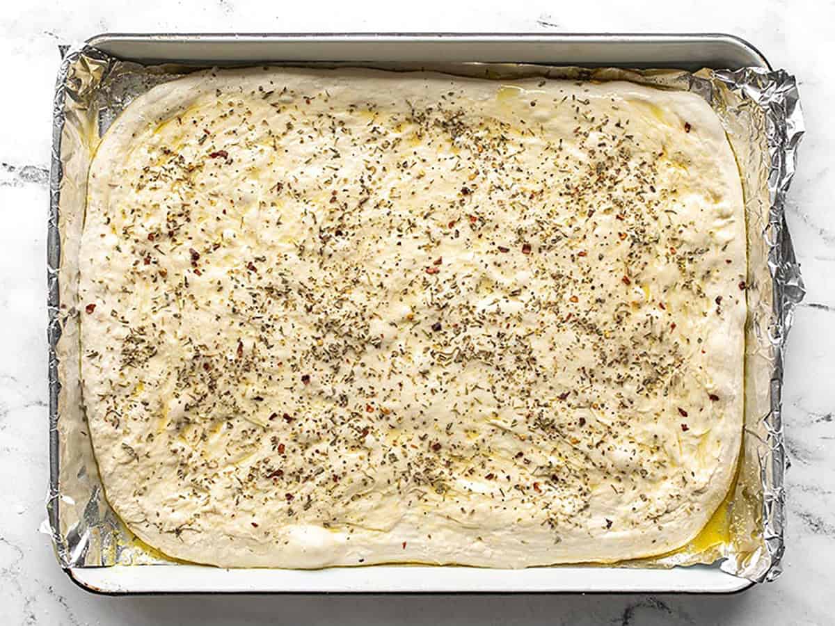 Focaccia dough stretched out onto the baking sheet and coated with herbs.