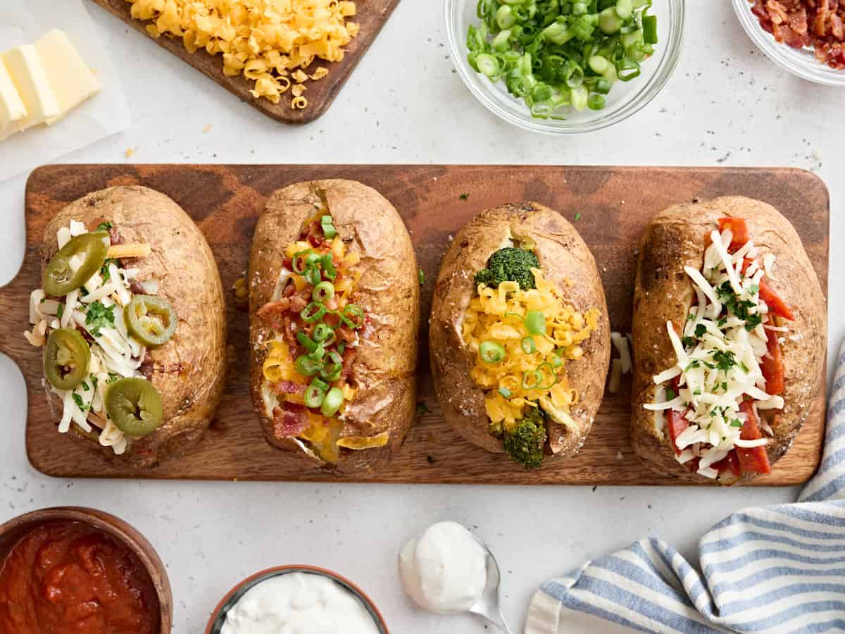 Four baked potatoes topped with various topping ideas and cheese on a wooden board.