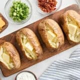Overhead view of four homemade baked potatoes on a wooden board, topped with butter.