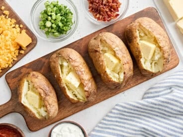 Overhead view of four homemade baked potatoes on a wooden board, topped with butter.