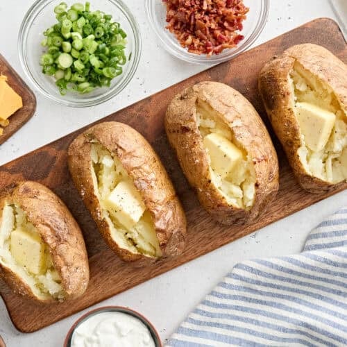 Overhead view of four homemade baked potatoes on a wooden board, topped with butter.