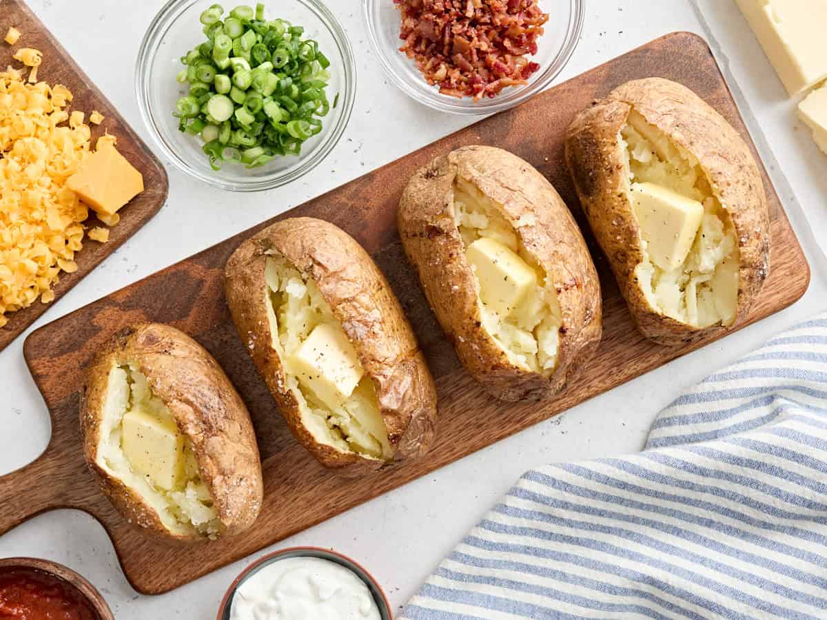 Overhead view of four homemade baked potatoes on a wooden board, topped with butter.