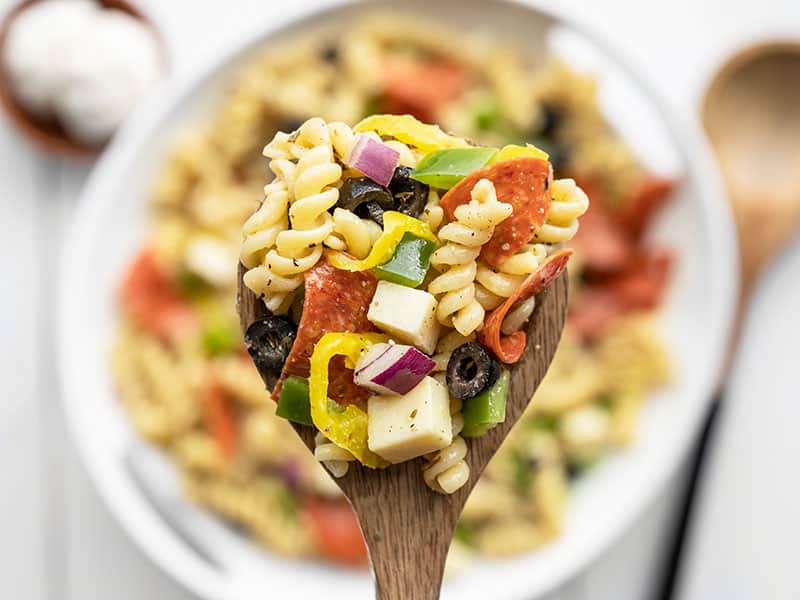 Repurpose your leftover pizza toppings into this deliciously simple Italian style pizza pasta salad. Including a simple homemade Italian dressing! Budetbytes.com Close up of a large spoonful of pizza pasta salad on a wooden spoon, bowl in the background