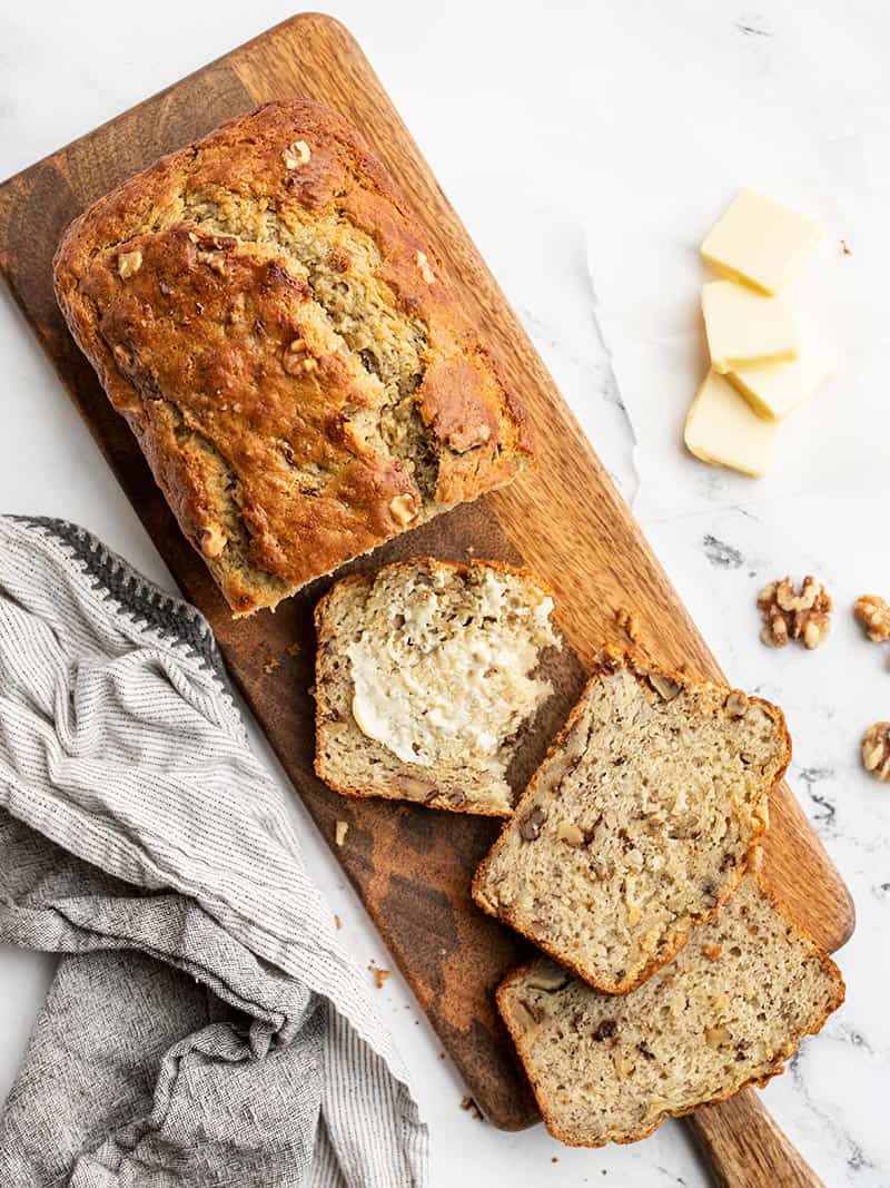 This super moist banana bread recipe uses plain yogurt to keep the bread soft and tender with less butter or oil. Add walnuts or even chocolate! BudgetBytes.com A loaf of banana bread on a narrow wooden cutting board, a few slices cut from the loaf and butter spread on one slice.