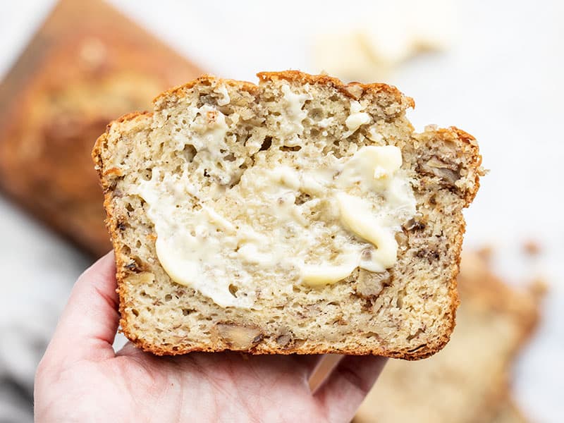 This super moist banana bread recipe uses plain yogurt to keep the bread soft and tender with less butter or oil. Add walnuts or even chocolate! BudgetBytes.com One slice of yogurt banana bread smeared with butter held close to the camera